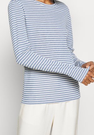 Personne portant une chemise à manches longues rayée bleu clair et blanc avec un pantalon couleur crème, ajustant la manche sur un fond clair.