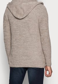 Beige gebreide hoodie van textuurstof. Heeft een ruime capuchon, lange mouwen en een rechte zoom. Minimale naden, casual ontwerp.