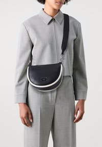 Bolso bandolera de cuero negro con forma curva, que presenta un acento blanco y una placa con el logotipo. Se lleva con un conjunto de patrón a cuadros.