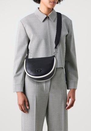 Zwarte leren schoudertas met een gebogen vorm, voorzien van een witte accent en een logo-plaatje. Draagt bij een outfit met een geruit patroon.