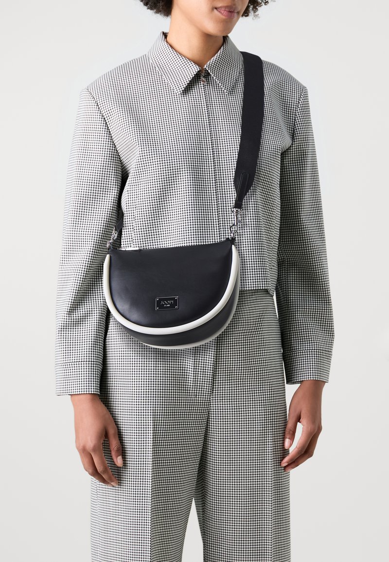 Bolso bandolera de cuero negro con forma curva, que presenta un acento blanco y una placa con el logotipo. Se lleva con un conjunto de patrón a cuadros.