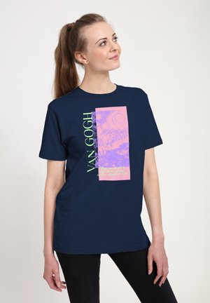 Femme aux cheveux bruns attachés en queue de cheval porte un t-shirt graphique Van Gogh bleu marine et un pantalon noir, regardant légèrement vers le haut avec un sourire.