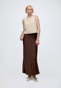 Femme aux cheveux blonds courts portant un haut beige texturé sans manches, une longue jupe marron, des talons beiges, et tenant une pochette beige.