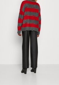 Person som bär en röd och grå randig stickad tröja, svarta vida läderbyxor och svarta klackade ankelskor, stående mot en enfärgad bakgrund.