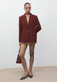 Vrouw in een bordeauxrode blazer, bruine minirok, zwarte puntige slingback hakken, parelketting, creolen en met een bijpassende bordeauxrode handtas.