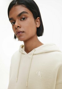 Sudadera color crema con cordones y logotipo bordado 'CK' en el pecho. Tela texturizada y suave. Diseño minimalista y tono neutro.