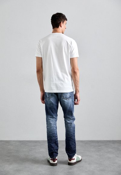 White short-sleeve t-shirt, blue faded jeans with subtle whiskering and contrast stitching, green and white striped shoes with a burgundy accent.