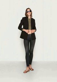 Sieviete, kas valkā melnu žaketi, olīvu krāsas īso topu, tumšas šauras bikses, brūnus sandales un melnas saulesbrilles, stāvot pret vienkāršu gaiši krāsotu fonu.