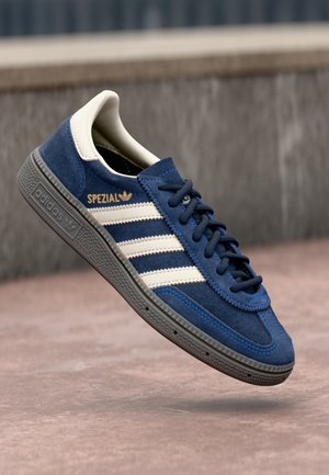 Blauer Adidas Spezial Wildledersneaker mit weißen Streifen und Gummisohle, schwebt in der Luft vor einem verschwommenen städtischen Hintergrund.