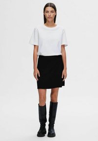 T-shirt bianco a maniche corte abbinato a una gonna nera, con un taglio dritto. Il modello indossa stivali neri fino al ginocchio. Design minimalista.