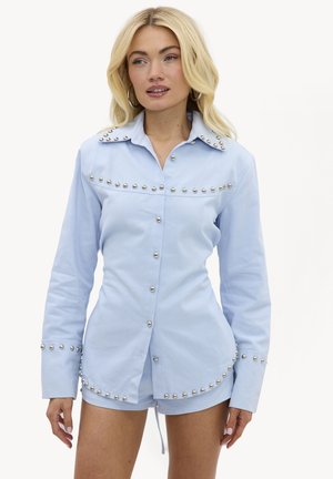 Femme blonde vêtue d'une chemise cloutée bleu clair à manches longues et d'un short assorti, debout devant un fond blanc uni.