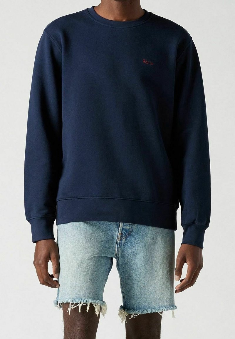 Personne portant un sweat-shirt bleu marine et un short en jean déchiré bleu clair, debout devant un fond clair uni.