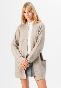 Beige gestrickte Strickjacke mit langen Ärmeln, offenem Schnitt und zwei seitlichen Taschen. Weiche Textur, geripptes Muster, mittellanges Design.