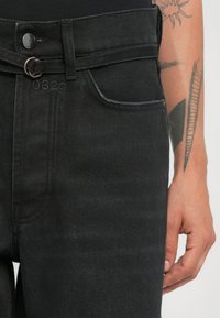 Close-up van zwarte spijkerbroek met een knoop, riemlus en een hand die naast een getatoeëerde onderarm rust.