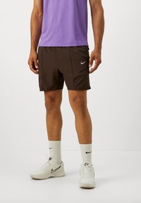 Man klädd i bruna Nike-shorts, vita Nike-strumpor, vita Nike-sneakers och en lila skjorta, stående mot en vit bakgrund.