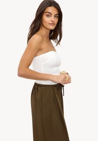 Jonge vrouw met lang, donker haar, gekleed in een witte strapless top en een hooggerokte olijfgroene rok, staand in profiel tegen een witte achtergrond.