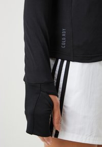 Schwarzes Langarmoberteil aus glattem Stoff mit Daumenlöchern in den Ärmeln und „COLD.RDY“-Text an der Seite, getragen über weißen Shorts mit Streifen.