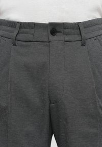 Pantaloni grigi strutturati con bottoni neri e passanti per la cintura, indossati con una camicia bianca a coste infilata, mostrati all'altezza della vita.