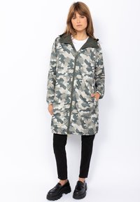 Giacca con motivo mimetico nelle tonalità di grigio e verde, dotata di zip frontale, cappuccio e due tasche laterali. Abbinata a pantaloni neri e scarpe.