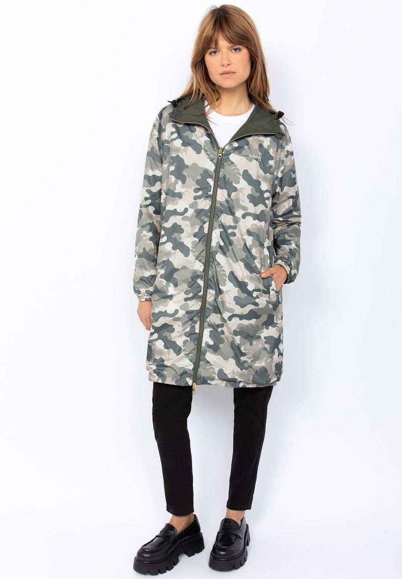 Veste à motif camouflage dans des teintes de gris et de vert, dotée d'une fermeture éclair à l'avant, d'une capuche et de deux poches latérales. Associée à un pantalon noir et des chaussures.