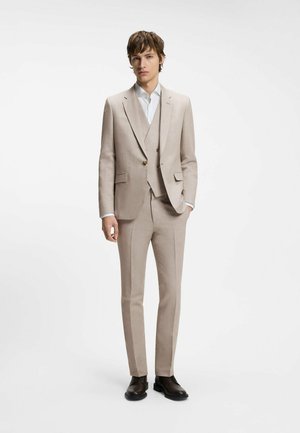 Giovane uomo che indossa un completo beige a tre pezzi con camicia bianca e scarpe eleganti nere, in piedi con una mano in tasca.