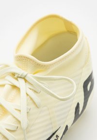 Zapatilla deportiva de color amarillo claro con diseño de caña alta, tejido texturizado, acentos en negro contrastante y cordones planos.