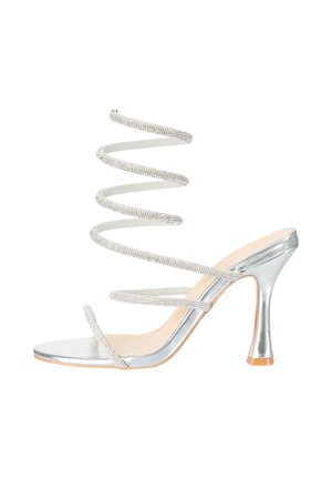 Sandalia plateada de tacón alto con correa espiral para el tobillo adornada con piedras brillantes y dos finas correas para los dedos también con piedras brillantes, sobre fondo blanco.