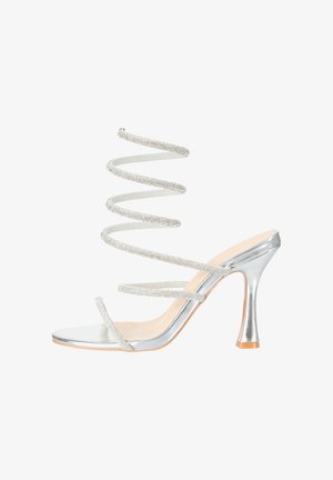 Sandalia plateada de tacón alto con correa espiral para el tobillo adornada con piedras brillantes y dos finas correas para los dedos también con piedras brillantes, sobre fondo blanco.