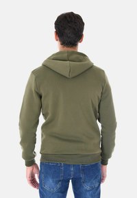 Felpa verde oliva realizzata in morbido tessuto, con cappuccio regolabile tramite cordino, polsini a costine e orlo. Indossata con jeans blu di denim.