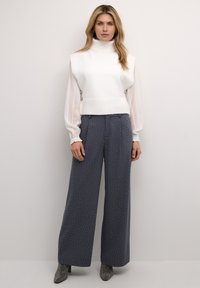 Biely pletený bezrukávový rolák cez priehľadnú blúzku s dlhým rukávom, doplnený tmavosivými nohavicami s širokými nohavicami a bielymi bodkami a striebornými akcentmi.