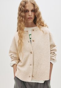 Svetlo béžový kardigán vyrobený z jemného materiálu, s okrúhlym výstrihom, zapínaním na gombíky a rebrovaným lemom. Modelka má na sebe vrstvený outfit.