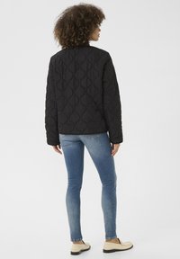 Zwarte gewatteerde jas met een gestructureerd patroon, gecombineerd met blauwe skinny jeans en lichtgekleurde loafers. Eenvoudig ontwerp, geen zichtbare sluitingen.