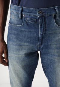 Denim kavbojke v različnih odtenkih modre barve s klasičnim pet-pocket dizajnom, ki vsebujejo gumbasto zapiranje in subtilne podrobnosti bledenja.