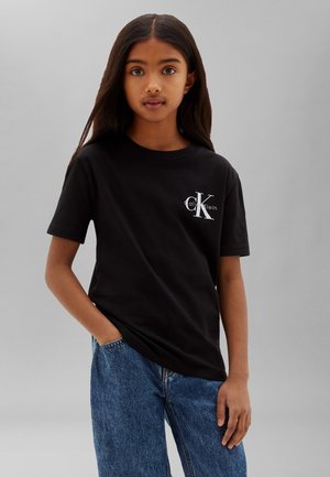 Pige med langt mørkt hår, iført en sort Calvin Klein T-shirt og blå jeans, stående med den ene hånd i lommen mod en grå baggrund.
