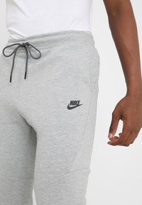 Närbild av en person som bär ljusgrå Nike joggingbyxor med dragsko i midjan och en vit t-shirt, med handen avslappnad vid sin sida.