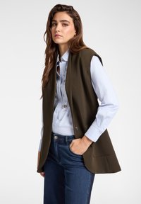 Olivově zelená bezrukávová vesta přes svetřík světle modré barvy s dekorativními ozdobami, doplněná tmavě modrými džíny. Hladká tkanina, strukturovaný design.