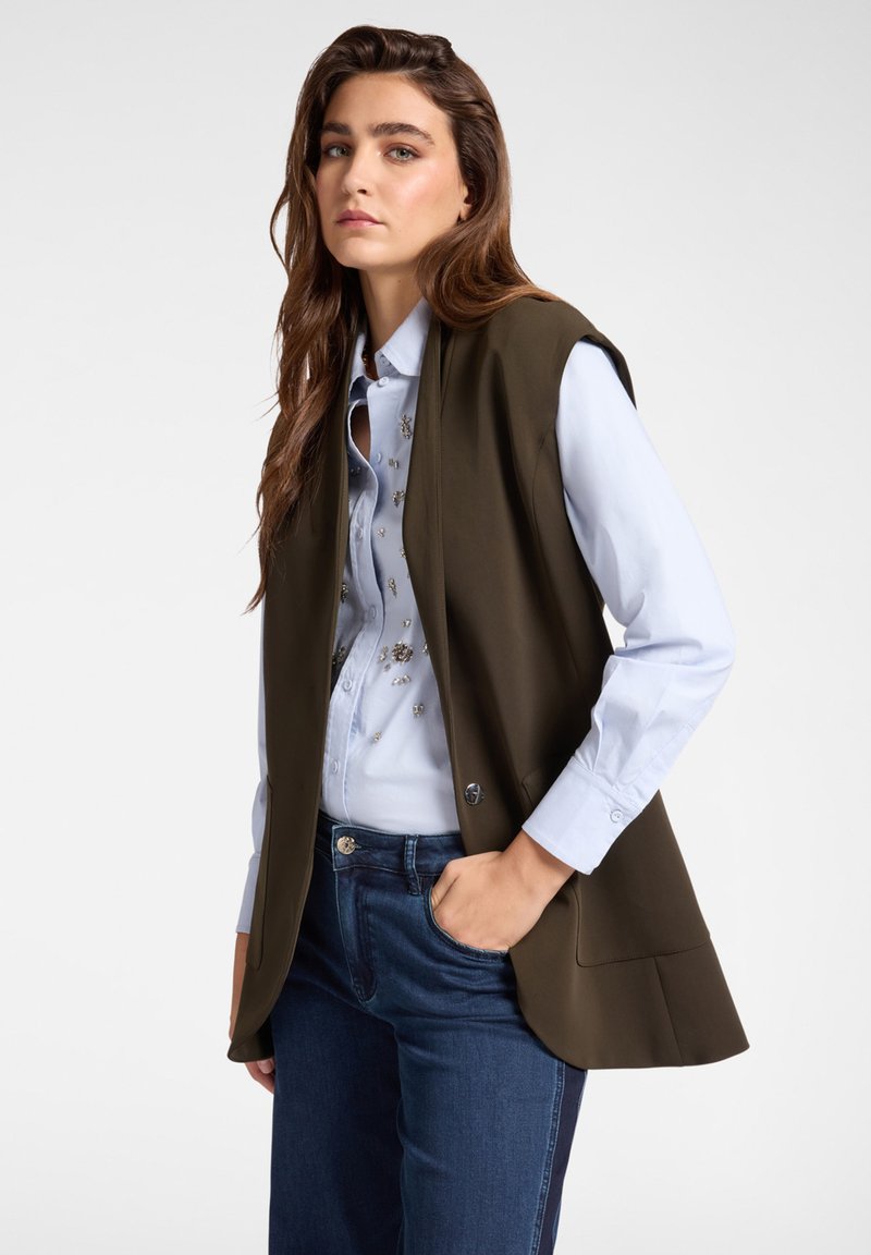 Olivově zelená bezrukávová vesta přes svetřík světle modré barvy s dekorativními ozdobami, doplněná tmavě modrými džíny. Hladká tkanina, strukturovaný design.