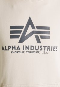 Felpa beige con logo stampato grigio scuro, caratterizzato da lettere stilizzate e strisce orizzontali, il testo include "ALPHA INDUSTRIES" e dettagli sulla posizione.