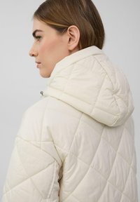 Gepolsterte cremefarbene Jacke mit einem rautenförmigen Steppmuster, einer verstellbaren Kapuze und einer weichen Textur, von der Seite dargestellt.