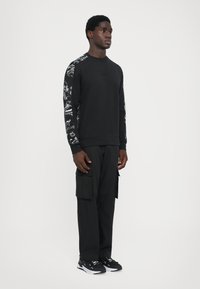 Sweatshirt preto com mangas estampadas, gola redonda e punhos canelados, combinado com calças cargo pretas e ténis pretos. Textura suave em toda a peça.