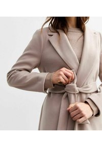 Ljusbeige bältad kappa i mjukt material med slagkrage och långa ärmar, med en jämn textur och enkel knyt-detalj i midjan.