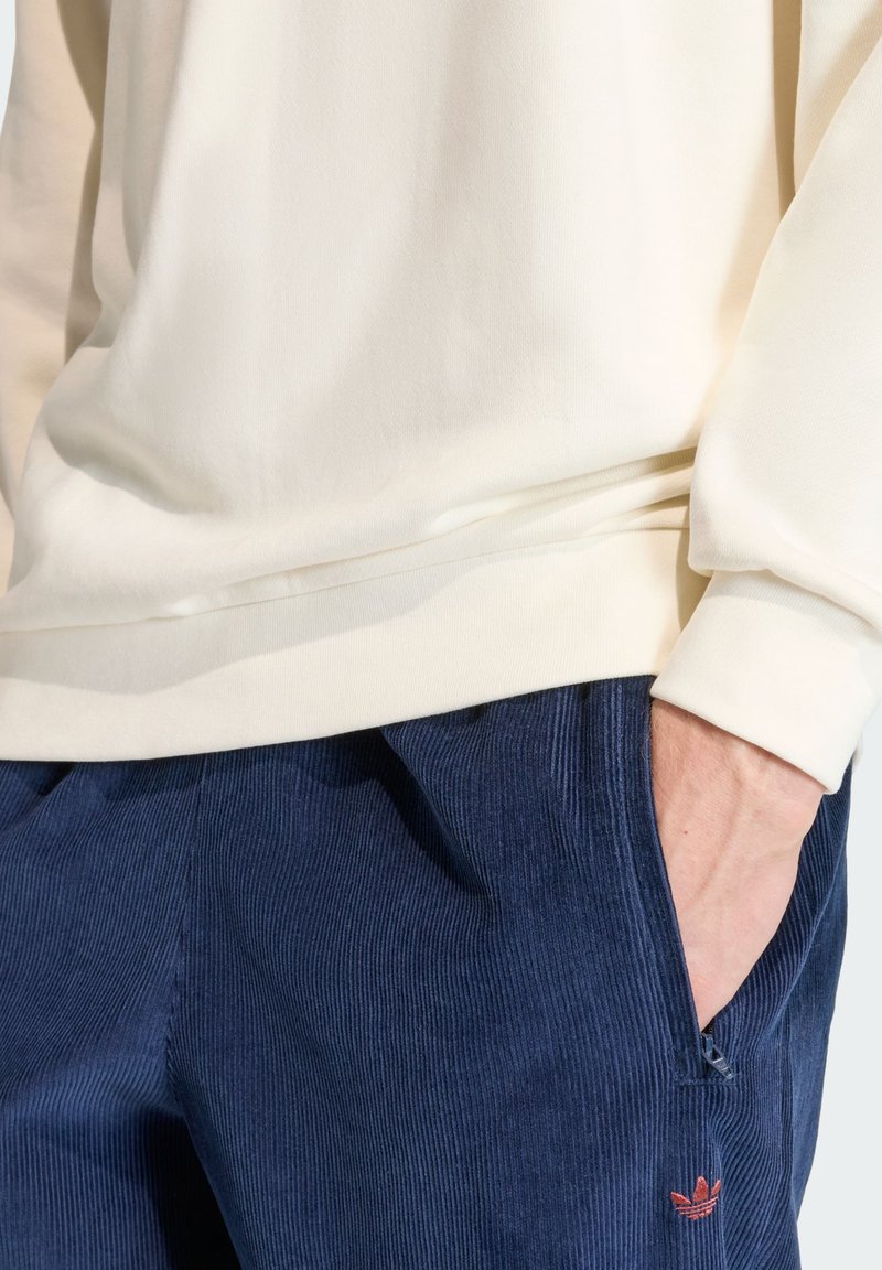 Sweat-shirt crème avec poignets côtelés associé à un pantalon en velours côtelé bleu foncé, avec un petit logo Adidas rouge brodé sur la poche.