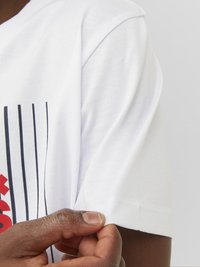 Camiseta blanca de algodón con mangas cortas, que presenta rayas verticales negras y un diseño rojo. Primer plano de una mano tocando la costura de la manga.