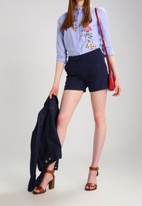 Blårandig skjorta med knappar och blommönster, marinblå shorts, bruna klacksandaler och en jeansjacka i handen. Röd axelväska.