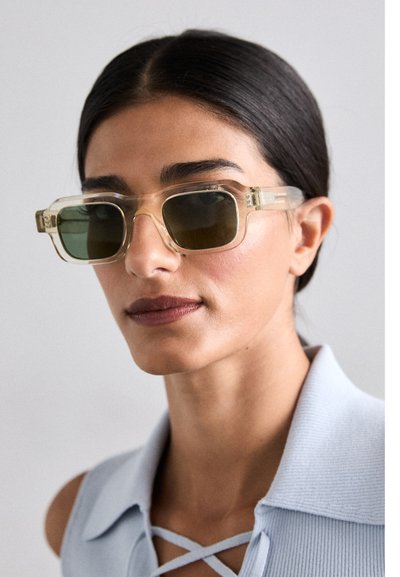 Gafas de sol cuadradas transparentes con lentes verdes oscuros. Las patillas presentan una sutil textura con un diseño de cierre. Se llevan con una blusa de color azul claro con cuello.