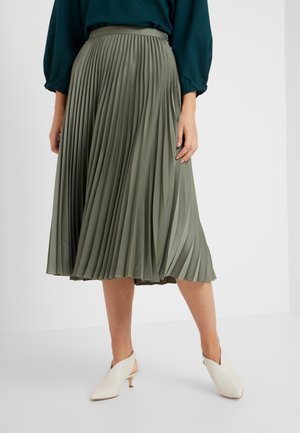 Kobieta ubrana w oliwkową plisowaną spódnicę midi, turkusowy długi rękaw na górze oraz białe buty na niskim obcasie typu slingback, stoi na białej podłodze.