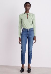 Lys grønn langermet topp med en nøkkelhulldetalj ved utringningen, kombinert med blå skinny jeans og svarte slip-on sko.