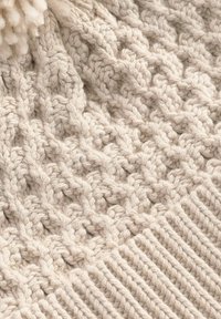 Cappello lavorato a maglia beige con un motivo a trecce testurizzato e bordo a coste. Presenta un pom-pom morbido sulla parte superiore, realizzato in filato soffice.