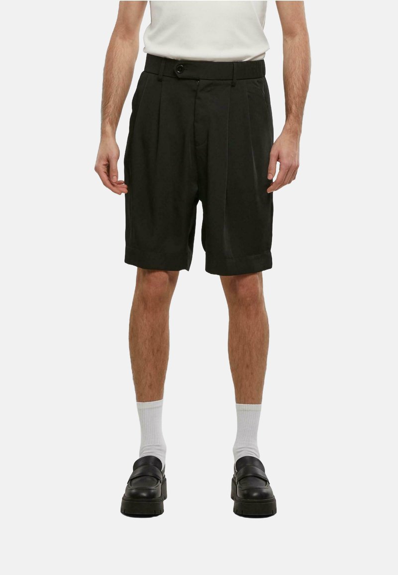 Svarte pregede shorts med knappeåpning, avslappet passform og skreddersydd stil. Brukt med hvite sokker og kraftige svarte sko.