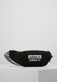 Černý plátěný ledvinka se zipem v hlavní přihrádce. Obsahuje bílé nášivky s logem Adidas a nastavovací popruh se značkovým opakovaným textem.